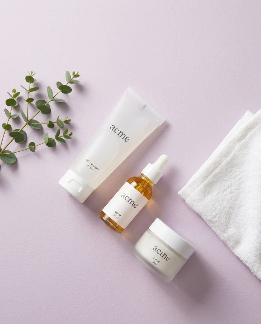 Flat lay of an Acme gel cleanser, serum and cream on lilac paper